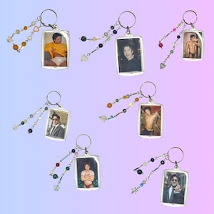 May include: Seven rectangular acrylic keychains, each featuring a unique photograph of a person. Every keychain is attached to a silver metal ring adorned with a dangling charm strand composed of various colorful beads, pearls, and star or heart shapes.