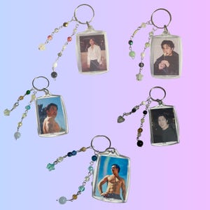 May include: Five clear acrylic keychains with silver key rings. Each keychain features a different photo of a person. The keychains are adorned with beaded strands in various colors, including blue, green, and pink.
