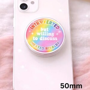 Op de afbeelding: Een witte smartphone met een ronde, kleurrijke pop-up grip bevestigd. De grip heeft de tekst "Introverted but willing to discuss Heated Rivalry" in een regenboogontwerp. De grip is 50 mm in diameter.