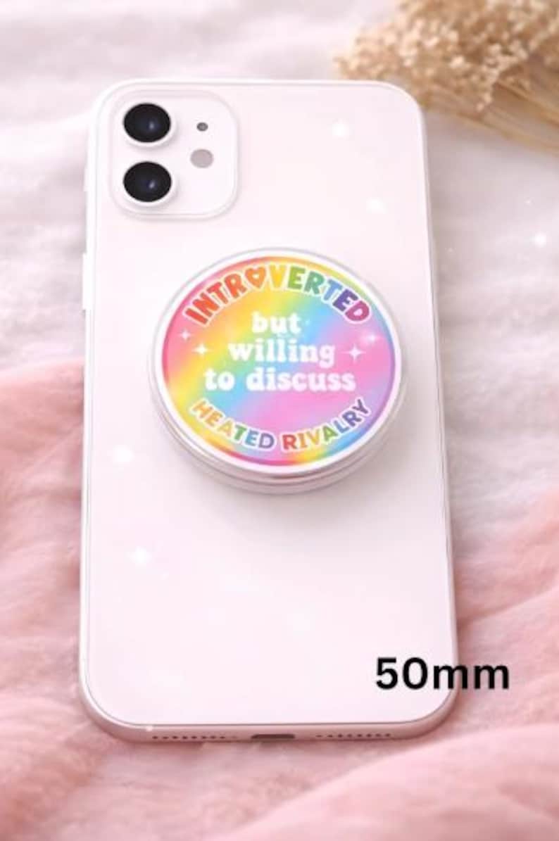 Op de afbeelding: Een witte smartphone met een ronde, kleurrijke pop-up grip bevestigd. De grip heeft de tekst "Introverted but willing to discuss Heated Rivalry" in een regenboogontwerp. De grip is 50 mm in diameter.