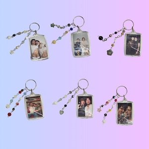 May include: A collection of six clear acrylic keychains, each featuring a different photograph. The keychains have silver-colored rings and are adorned with beaded chains in various colors, including white, clear, and colorful beads.