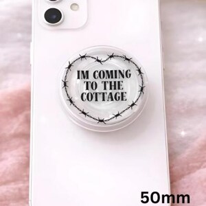Op de afbeelding: Een witte smartphone met een popsocket bevestigd. De popsokkel heeft een transparante achtergrond met een zwart prikkeldraad hart ontwerp en de tekst "IM COMING TO THE COTTAGE". De popsokkel is 5 cm in diameter.