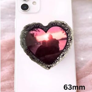 Op de afbeelding: Een witte smartphone met een hartvormige telefoonhouder. De houder heeft een zonsondergangfoto in een zwarte, kantachtige rand. De houder meet 6,3 cm.
