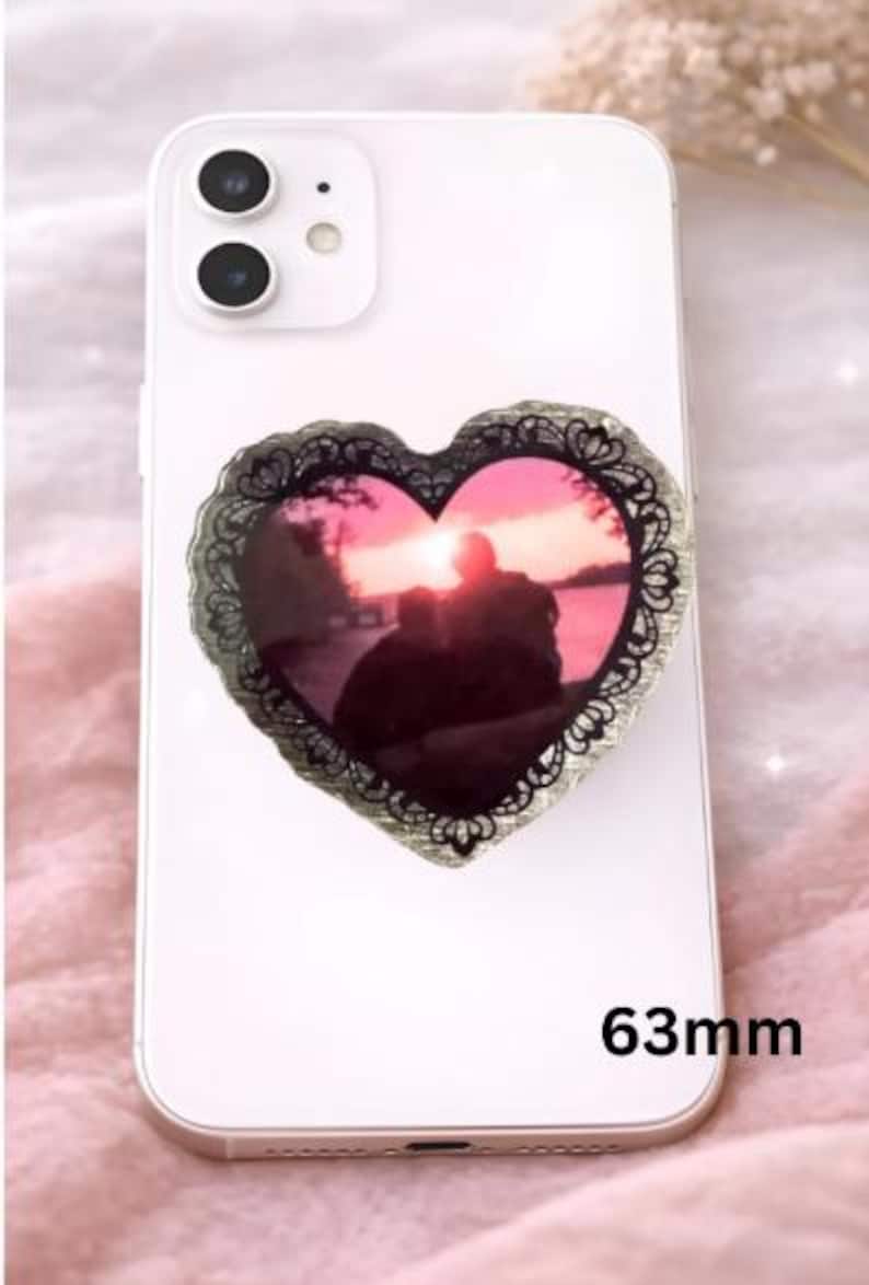 Op de afbeelding: Een witte smartphone met een hartvormige telefoonhouder. De houder heeft een zonsondergangfoto in een zwarte, kantachtige rand. De houder meet 6,3 cm.