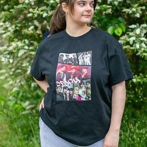 May include: Black t-shirt with a collage of images and the word "ORACHA" in red. The person is wearing the shirt outside, with green foliage behind them. The shirt is paired with grey sweatpants.