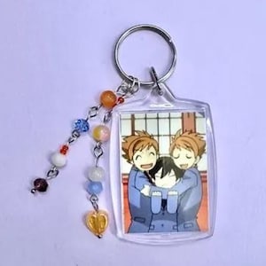 May include: A clear acrylic keychain featuring a colorful anime-style image of three characters hugging. The keychain has a silver metal ring and a beaded charm with orange, blue, and white beads, and a yellow heart-shaped charm.