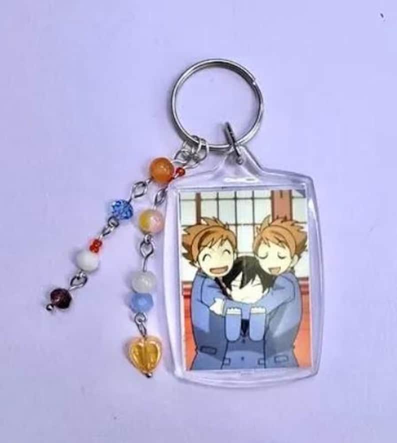 May include: A clear acrylic keychain featuring a colorful anime-style image of three characters hugging. The keychain has a silver metal ring and a beaded charm with orange, blue, and white beads, and a yellow heart-shaped charm.