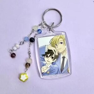 May include: A rectangular clear acrylic keychain featuring an anime image of two male characters. The keychain has a silver ring and a beaded charm with white, blue, black, and brown beads, and a yellow flower charm.