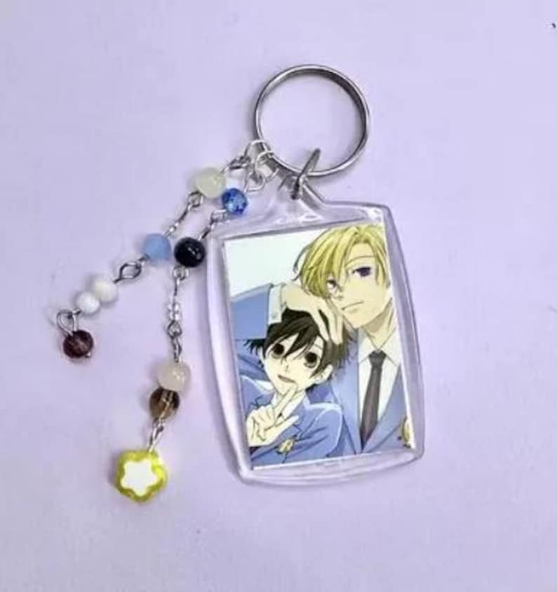 May include: A rectangular clear acrylic keychain featuring an anime image of two male characters. The keychain has a silver ring and a beaded charm with white, blue, black, and brown beads, and a yellow flower charm.