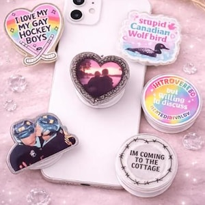 Op de afbeelding: Een verzameling telefoonhouders met diverse ontwerpen en tekst, waaronder een hart met de tekst "I LOVE MY GAY HOCKEY BOYS", een hart met een foto en een ronde houder met de tekst "IM COMING TO THE COTTAGE".