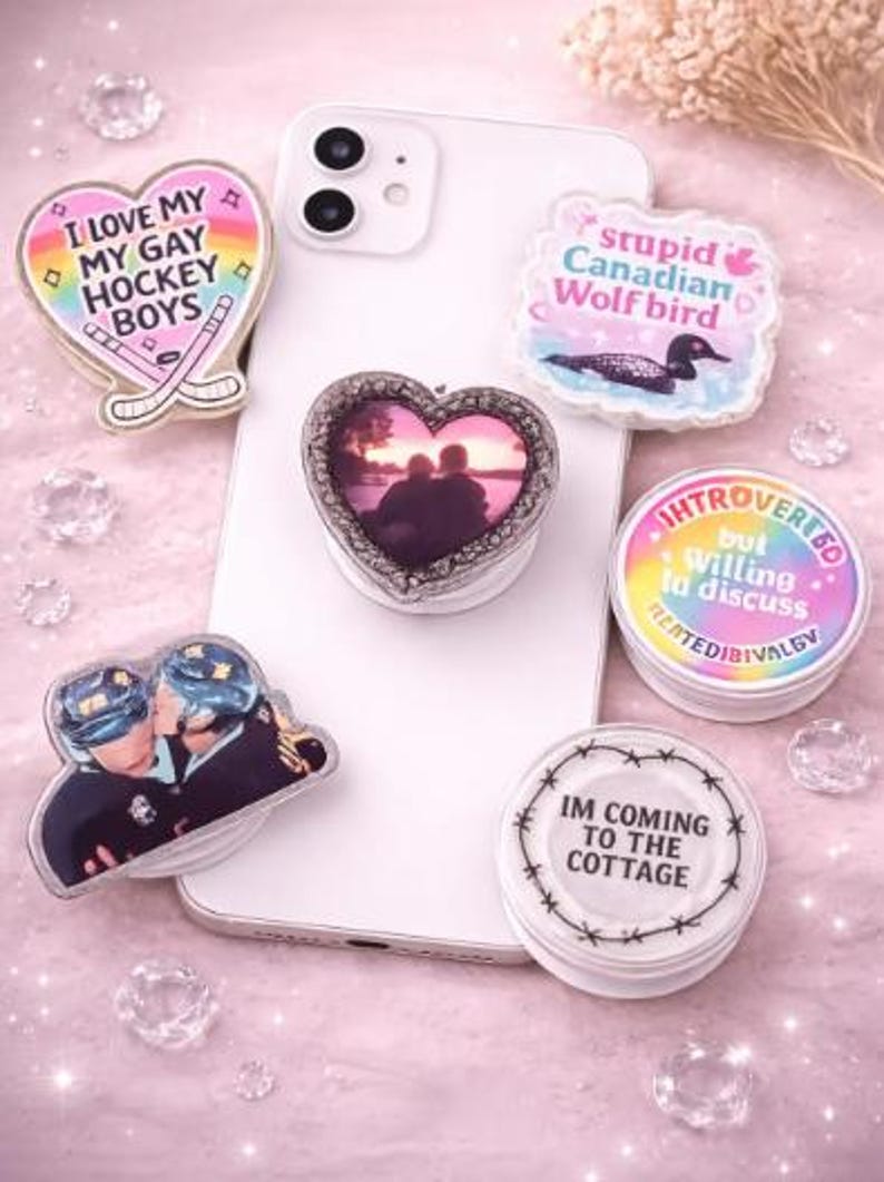 Op de afbeelding: Een verzameling telefoonhouders met diverse ontwerpen en tekst, waaronder een hart met de tekst "I LOVE MY GAY HOCKEY BOYS", een hart met een foto en een ronde houder met de tekst "IM COMING TO THE COTTAGE".