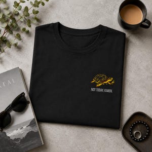 Könnte beinhalten: Ein schwarzes T-Shirt mit einer gestickten gelben Schildkröte auf der rechten Seite und dem darunter in Weiß gedruckten Text "NOT TODAY, KAREN.". Das T-Shirt ist ordentlich gefaltet und liegt zusammen mit verschiedenen Accessoires auf einer Fläche.