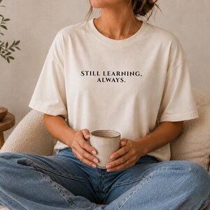 Könnte beinhalten: Ein elfenbeinfarbenes Kurzarm-T-Shirt mit dem schwarzen Schriftzug "STILL LEARNING, ALWAYS." in einer Serifenschrift auf der Brust. Das Kleidungsstück hat einen Rundhalsausschnitt und eine lockere Passform, kombiniert mit einer blauen Jeans.