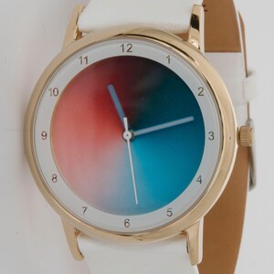May include: A gold-tone wristwatch with a white leather band. The watch face features a gradient of red, blue, and white. The watch has a gold bezel and white hands. The watch band is white and has a buckle closure.
