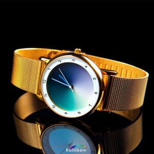 May include: Gold-toned wristwatch with a gradient blue and green face and a gold mesh band. The watch face has white numerals and hands. The watch is displayed on a reflective black surface.