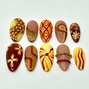 May include: A set of ten almond-shaped press-on nails featuring a warm color palette of brown, orange, yellow, and cream. The designs include intricate patterns such as polka dots, swirling lines, floral motifs, and embossed textures with gold accents.