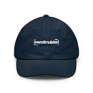 May include: A navy blue baseball cap featuring the word overstimulated embroidered in white lowercase letters across the front center. The text is flanked by small white horizontal lines, and the cap has a curved brim and eyelets.