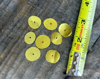 May include: A collection of small, round, gold-colored metal discs with a hole in the center, scattered on a weathered wooden surface. A yellow Stanley measuring tape is positioned next to the discs, showing measurements in inches.