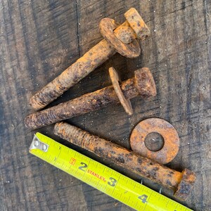 Op de afbeelding: Close-up van drie roestige bouten met ringen en een gele meetlint. De bouten zijn ongeveer 12.7 cm lang. De items liggen op een verweerd houten oppervlak.