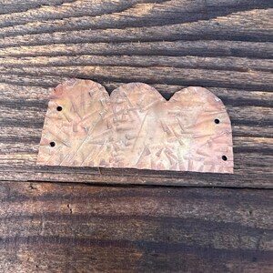 May include: A copper-coloured metal piece with a textured surface and a scalloped edge. The piece has two holes on each side.