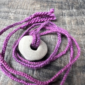 May include: A purple braided cord with a gray stone tied in the center. The cord is looped and the stone is in the center of the loop.