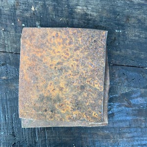 Puede incluir: Una pila de láminas cuadradas de metal oxidado con una superficie moteada de color naranja y marrón. Las láminas están apiladas unas sobre otras, con los bordes ligeramente irregulares. El fondo es una superficie de madera oscura y desgastada.
