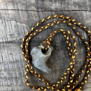 May include: A gray stone pendant with a unique shape is suspended from a braided cord necklace. The cord is brown with yellow accents. The necklace is displayed on a weathered wooden surface.