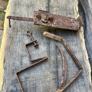 May include: A collection of antique, rusty metal hardware pieces. Includes a latch, a clamp, and several bent metal rods. The items are arranged on a weathered wooden surface, showcasing their aged appearance and historical significance.
