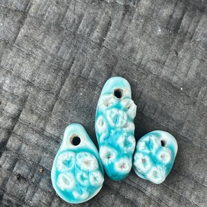 Pode incluir: Três pingentes de cerâmica feitos à mão em tons de turquesa e branco. Cada pingente tem uma forma e textura únicas, com um furo na parte superior para enfiar. Os pingentes são exibidos em uma superfície de madeira envelhecida.