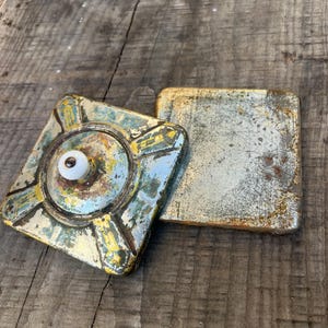 Könnte beinhalten: Zwei quadratische Vintage-Aschenbecher mit abgenutzter Farbe. Ein Aschenbecher hat einen weißen Knopf und ein kreisförmiges Design mit gelben und blauen Akzenten. Der andere Aschenbecher ist ein schlichtes Quadrat mit Metallic-Finish. Beide liegen auf einer verwitterten Holzoberfläche.