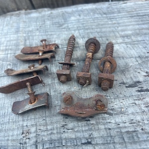 Könnte beinhalten: Eine Ansammlung von verwitterten, rostigen Metallbeschlägen, bestehend aus mehreren Gewindebolzen mit Vierkantmuttern und Unterlegscheiben sowie mehreren gebogenen Metallmontagewinkeln mit kleinen, daran befestigten Schrauben.