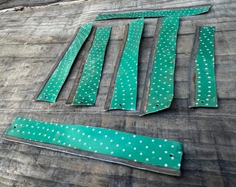 May include: A collection of green metal strips with white polka dots, arranged on a weathered wooden surface. The strips vary in size and shape, some with small holes. The overall aesthetic is vintage or repurposed.