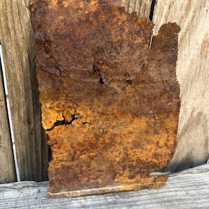May include: A rectangular sheet of metal with heavy oxidation and rust, displaying a deep orange and brown color palette. The surface is weathered and textured with visible corrosion, jagged edges, and several small holes throughout the material.