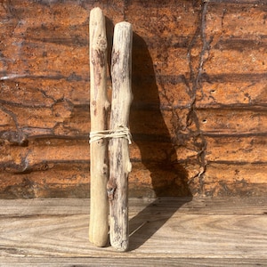 May include: Two weathered driftwood pieces tied together with natural twine. The wood has a light brown color with darker knots and grain patterns. The background is a rusty corrugated metal sheet.