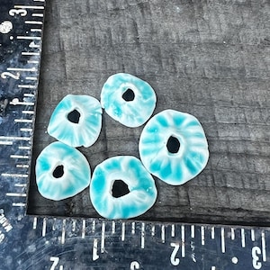 Pode incluir: Seis contas artesanais em forma de donut com um padrão de redemoinho turquesa e branco. Cada conta tem um buraco central. As contas estão dispostas sobre uma superfície de madeira escura, perto de uma régua.