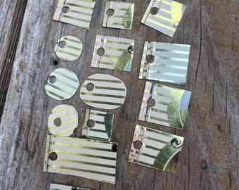 May include: A collection of small, metallic pieces with a gold and white striped pattern. The pieces are various shapes, including rectangles, squares, and circles, and have small holes. They are scattered on a weathered wooden surface.