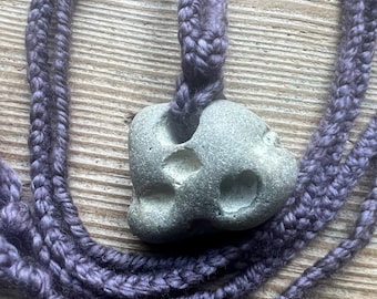 May include: A gray stone pendant with a unique shape and texture, hanging from a purple braided cord.