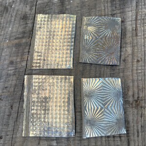 Może przedstawiać: Cztery kwadratowe metalowe płytki o teksturowanych i wzorzystych powierzchniach. Dwie płytki mają wzór kratki, a pozostałe dwa wzór rozbłysku gwiazdy. Płytki mają metaliczny, postarzany wygląd z różnymi odcieniami.