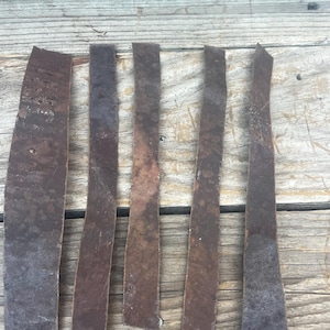 May include: Five long, narrow strips of dark brown leather with a matte, textured surface. Each strip features irregular edges and a weathered appearance, showing variations in tone and surface markings across their length.