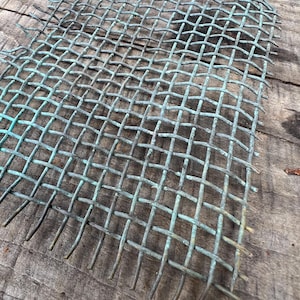 May include: A weathered, square-patterned metal mesh with a greenish-blue patina. The mesh is made of thin, intersecting wires, creating a grid-like structure. The edges of the mesh have exposed wire ends. The mesh is placed on a rough, wooden surface.
