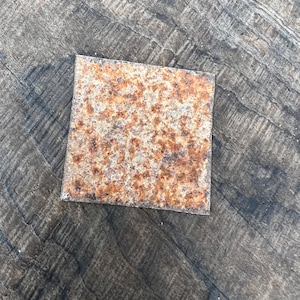 May include: A square piece of rusty metal with a rough, textured surface. The metal is a mix of brown and orange hues, showing signs of wear and age.