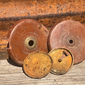 Op de afbeelding: Twee antieke bruin lederen meetwielen met messing centra en twee losgemaakte, verroeste metalen deksels. De wielen zijn cirkelvormig en vertonen tekenen van slijtage en ouderdom. De achtergrond is verweerd, gegolfd metaal.