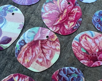 May include: A collection of floral-patterned, circular tags in various sizes. The tags feature vibrant blooms in shades of purple, pink, and blue, with small holes for stringing. The tags are arranged on a textured, dark gray surface.