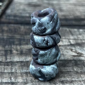 Puede incluir: Una pila de cuatro cuentas de cerámica hechas a mano. Las cuentas son una mezcla de gris y marrón, con una apariencia texturizada y orgánica. Cada cuenta tiene aproximadamente 2,5 cm de diámetro y están apiladas una encima de la otra.