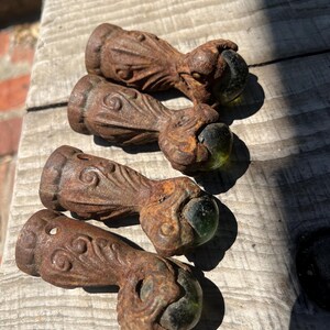 May include: Four antique furniture feet featuring a cast iron claw and ball design with a rusted brown finish. Each piece displays intricate scrollwork on the leg and holds a dark green glass sphere within the metal talons of the claw.