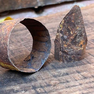 Puede incluir: Primer plano de dos piezas de metal oxidado. Una es una banda curva, la otra es una pieza triangular. El metal tiene una superficie rugosa y texturizada con tonos marrones y anaranjados. Los objetos están sobre una superficie de madera.