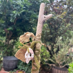 Peut inclure: Une branche de bois flotté brun clair enveloppée de fil de laine vert olive et d'une bande de tissu beige. Un petit coquillage en spirale rose pâle est attaché à la branche avec le fil vert, positionné près du centre du bâton décoratif.