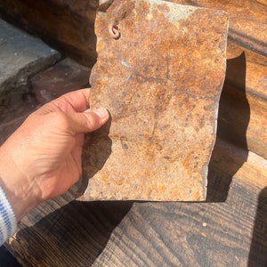 Puede incluir: Una lámina de metal oxidada con un agujero en la esquina superior izquierda. La lámina es marrón y naranja con una textura rugosa.