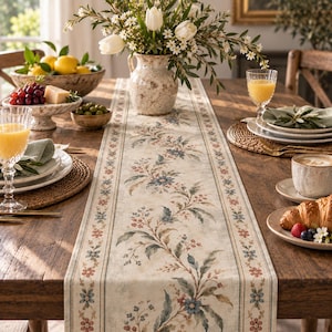 May include: A long rectangular table runner featuring a cream-colored background with a central botanical pattern of blue and red flowers, green leaves, and small berries, framed by thin decorative borders with repeating floral motifs.