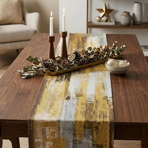 May include: A rectangular table runner featuring an abstract brushstroke pattern in shades of mustard yellow, white, and dark brown. The runner is displayed on a wooden table with two tall wooden candle holders, a gold tray, and pinecones.